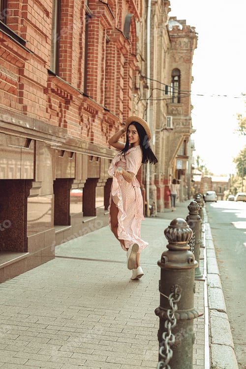 Preview: A young brunette in a soft pink dress with a hat walks around the city