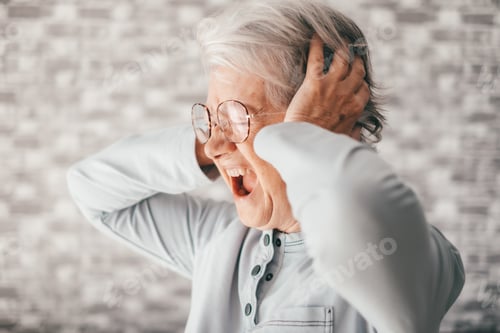 Preview: Desperate frustrated senior woman screaming in pain with hands on ears, sitting at home alone