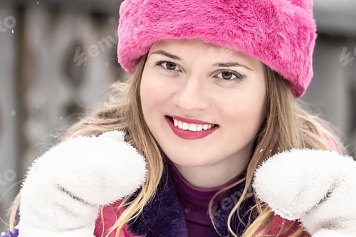 Vorschau: eine lächelnde junge Frau mit rosafarbenem Schal und Hut, Fäustlingen und einem lila Pelzmantel draußen im Winter.