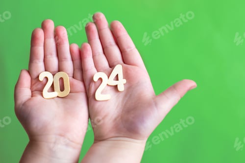 Preview: Child hands holding wooden number 2024 on palms on festive green background with copy space