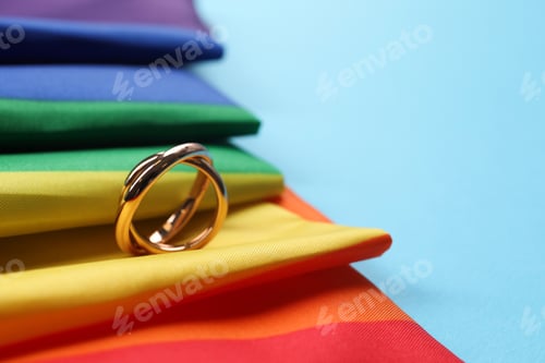 Preview: Rainbow LGBT flag and wedding rings on light blue background, closeup. Space for text