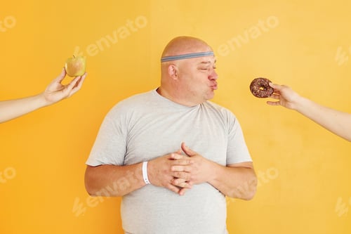 Preview: Apple or cookie. Funny overweight man in sportive head tie is against yellow background