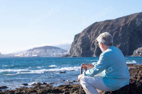 Preview: Back view of senior man sitting at sunset light on the beach holding a walking stick