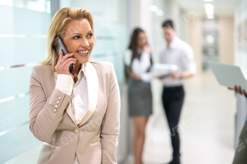 Preview: Portrait of middle aged blonde business woman in formalwear talking on mobile phone in office.