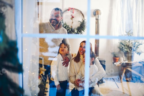 Preview: Christmas time. Happy family with two little kids gathered at window at home decorating xmas tree