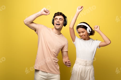 Preview: happy young man and woman dancing while listening music with headphones on yellow background