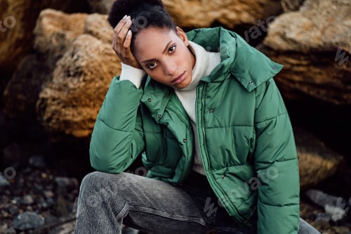 Preview: African American girl in stylish down jacket intently looking in camera posing on stones by the sea