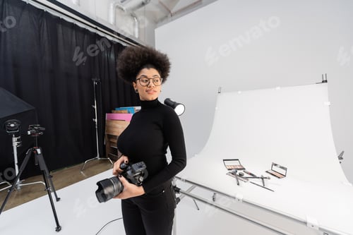 Preview: happy african american content maker with digital camera looking away near decorative cosmetics on