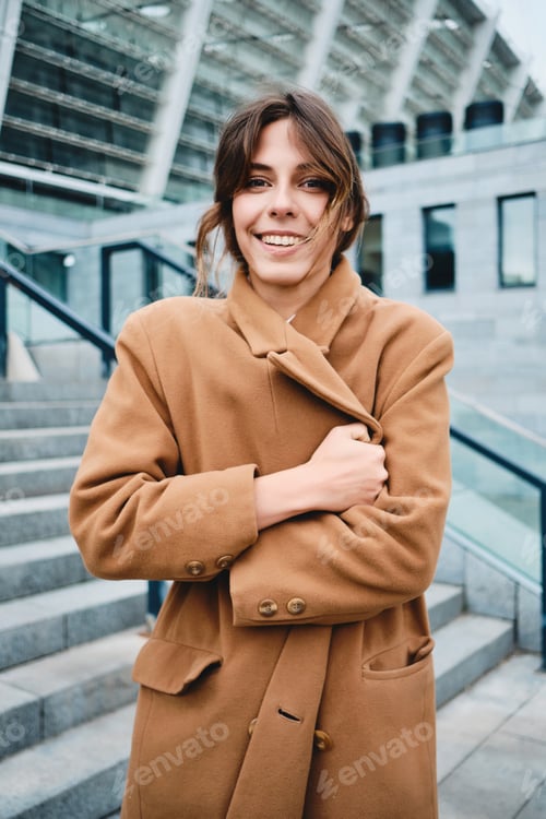 Preview: Young pretty smiling woman fastening up coat from cold happily looking in camera outdoor
