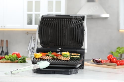 Preview: Electric grill with tasty meat and vegetables on white marble table in kitchen