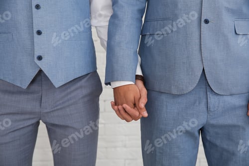 Preview: Newlywed gay couple holding hands near white wall, closeup