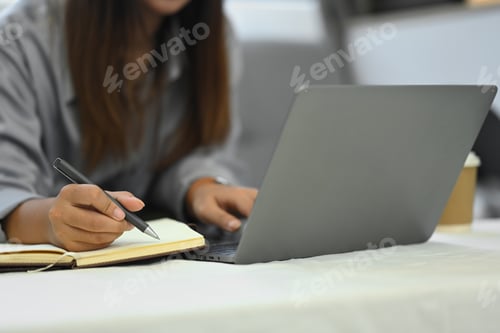 Preview: Young female freelancer working with laptop and writing in a notebook