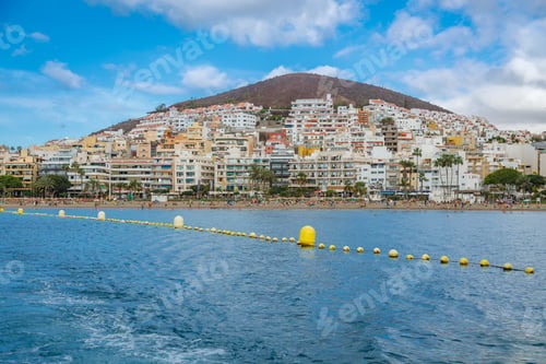 Preview: Panoramic view of Los Cristianos resort town with colorful buildings cascading up volcanic hillside