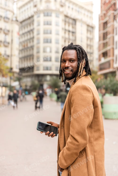 Preview: smiling african american man holds a mobile phone looking at camera walking on the street