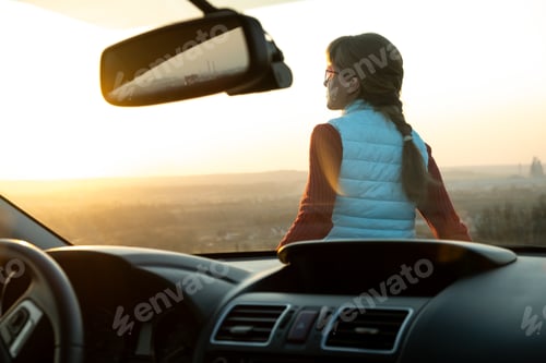Preview: View from inside of a young woman standing near her car enjoying warm sunset view.
