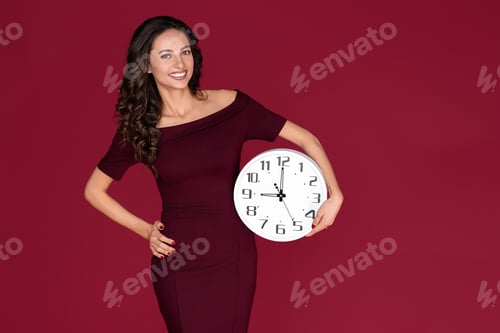 Preview: Happy young modern business woman in maroon dress holding a clock over red background