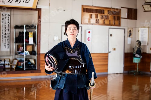 Preview: Female Japanese Kendo fighter standing in a gym, holding Kendo mask and sword, looking at camera.