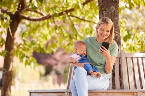 Preview: Mother Sitting On Park Bench Under Tree With Baby Daughter Looking At Mobile Phone