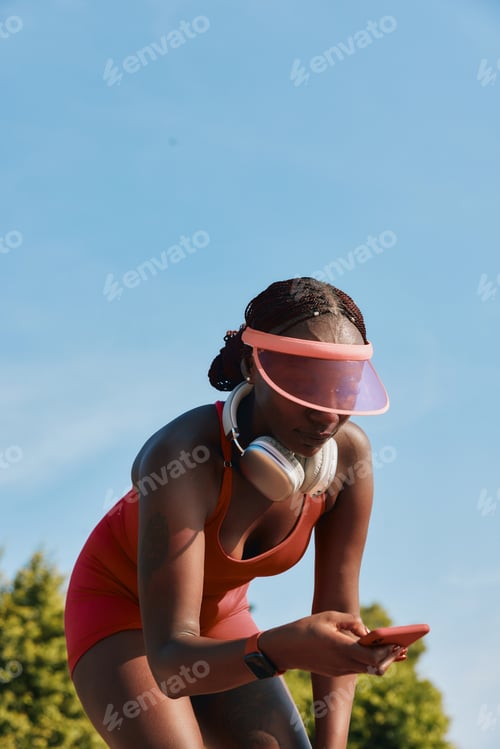 Preview: Young woman in activewear using smartphone outdoors, wearing headphones and a visor under clear blue