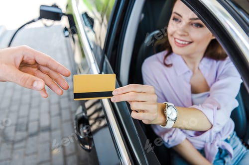 Preview: selective focus of cheerful woman sitting in car and giving credit card to worker at gas station