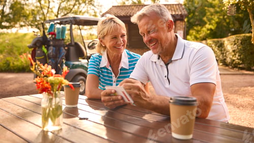Preview: Senior Couple Sitting Having Coffee After Round Of Golf Looking At Score Card Together