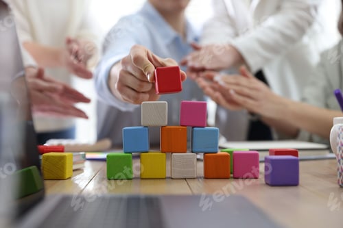 Preview: Unity concept. People building pyramid of colorful cubes at table indoors, closeup