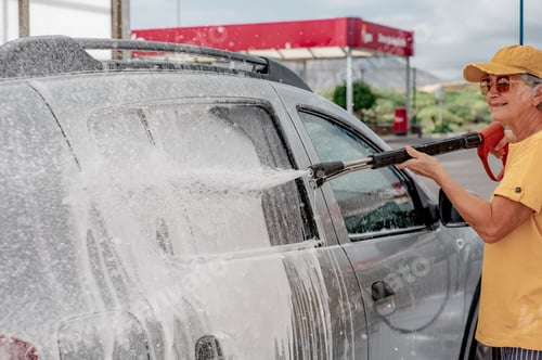 Preview: Smiling senior woman washing car in a self-service car wash station using high pressure water pump
