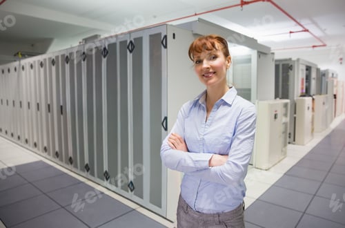 Preview: Female technician standing arms crossed in blue shirt gray pants at data center racks, copy space