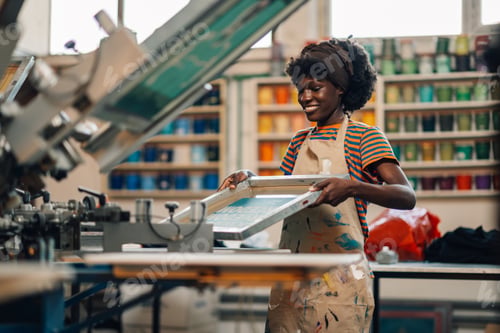 Preview: Happy multiracial press woman adjusting screen press for printing.