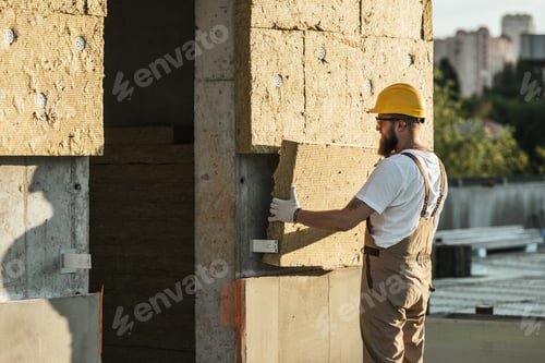 Preview: side view of builder in protective helmet and googles doing insulation of wall at construction site