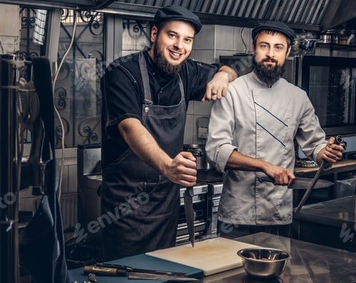 Preview: Two bearded cooks dressed in uniforms and hats posing in kitchen, looking at a camera.