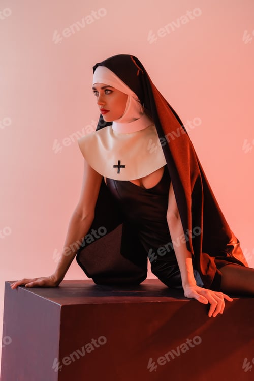 Preview: young nun in sexy dress looking away while sitting on black cube isolated on pink