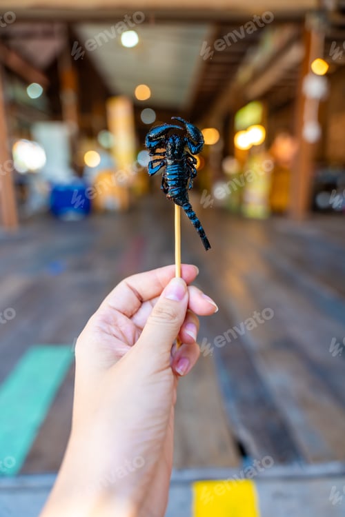 Preview: Fried scorpion on a skewer in hand of tourist in floating open air market on the pond in Pattaya