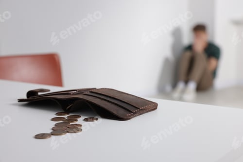 Preview: Upset man sitting on floor indoors, focus on wallet with coins. Financial problems