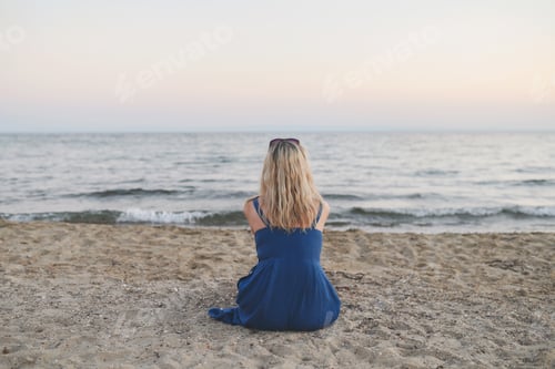 Preview: Beautiful young woman with blonde hair sits on beach and looking at the ocean. From the back view