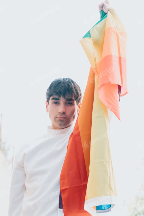 Preview: Portrait of young gay man with lgtbi lgbtq flag in the city on a summer day between the buildings