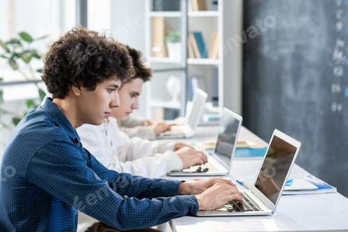 Preview: Side view of two guys sitting by desk in row in front of laptops