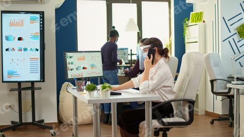 Preview: Freelancer with mask talking on mobile phone in fron of computer