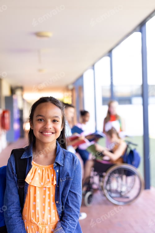 Preview: Portrait of smiling biracial schoolgirl standing with bag in school corridor