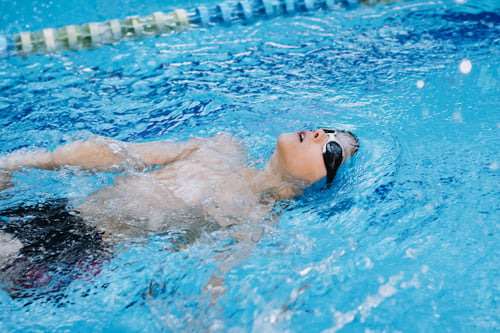 Preview: little caucasian boy wearing goggles swimming backstroke in a pool