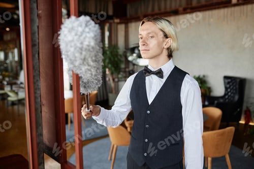 Preview: Elegant young man wearing bowtie cleaning with feather duster in restaurant hall
