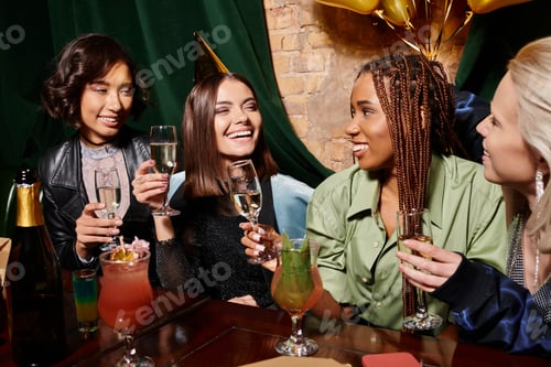 Preview: young birthday girl in party hat holding champagne near multiethnic girlfriends and cocktails in bar