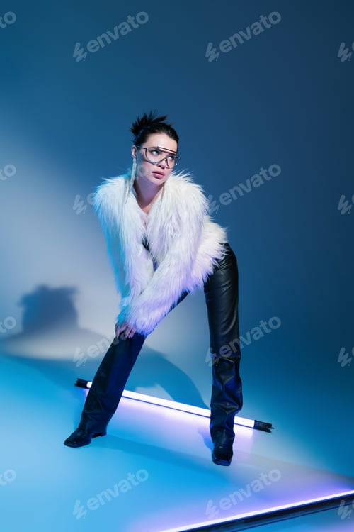 Preview: brunette model in black leather pants and white faux fur jacket looking away near fluorescent lamp
