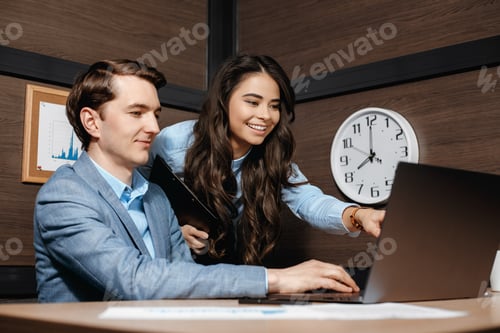 Preview: Two happy colleagues working together pointing at laptop and business documents in modern office.