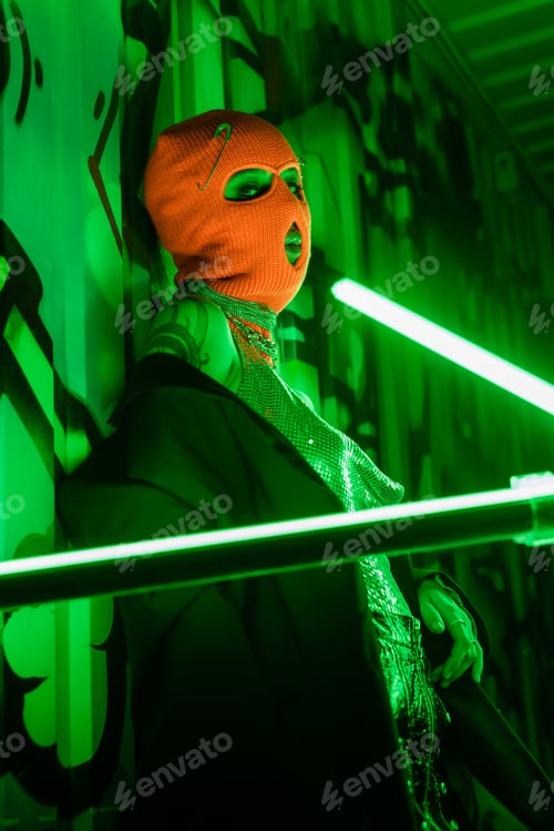 Preview: sexy woman in orange balaclava and black blazer looking at camera near bright neon lamps and green