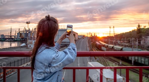 Preview: A young woman photographs a beautiful sunset from a bridge.