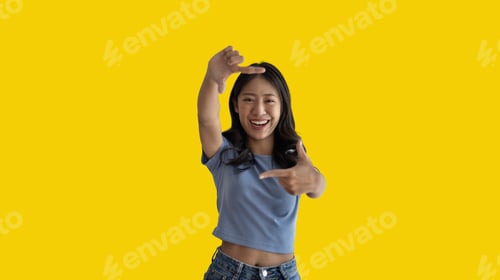 Vista previa: Joven asiática juguetona de buen humor posando para una foto o marco de fotos con los dedos sobre fondo amarillo