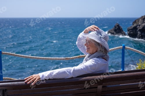 Preview: Smiling senior woman dressed in white sitting outdoors by the sea on a sunny day admiring the ocean