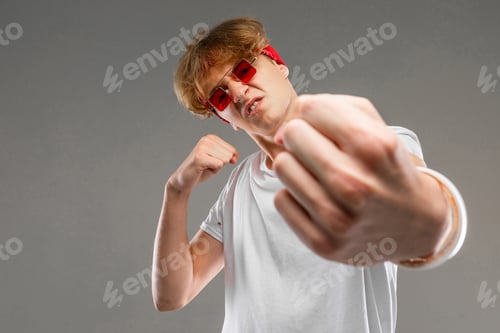 Preview: Young caucasian man in red sunglasses, white t-shirt posing on camera isolated on grey background