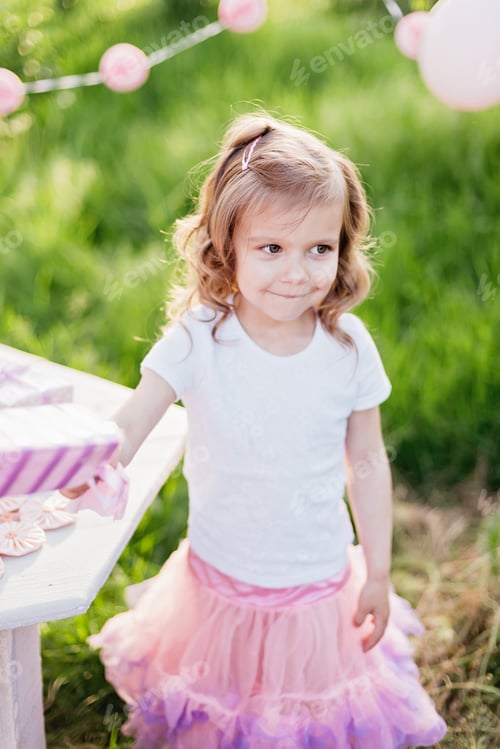 Preview: Happy birthday little girl making wish blowing candles on cake with pink decor in beautiful garden